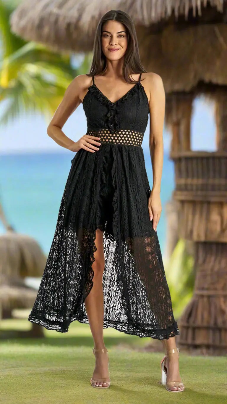 A woman smiles outdoors in a La Moda Clothing Lace Romper Beach Dress with Shorts, featuring a sleeveless black lace design, see-through midriff, high-low hem, and clear heels. Tropical huts, palm trees, and the ocean appear in the background.