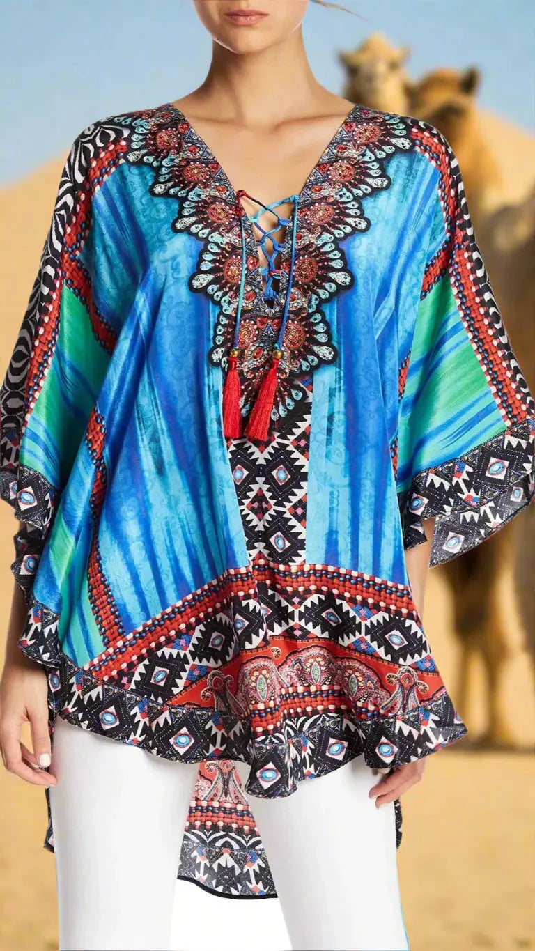 A woman models the La Moda Clothing Kaftan for Women, featuring vibrant blue, red, and black geometric and floral patterns with a lace-up neckline and wide sleeves, standing before desert dunes with blurred camels in the background.