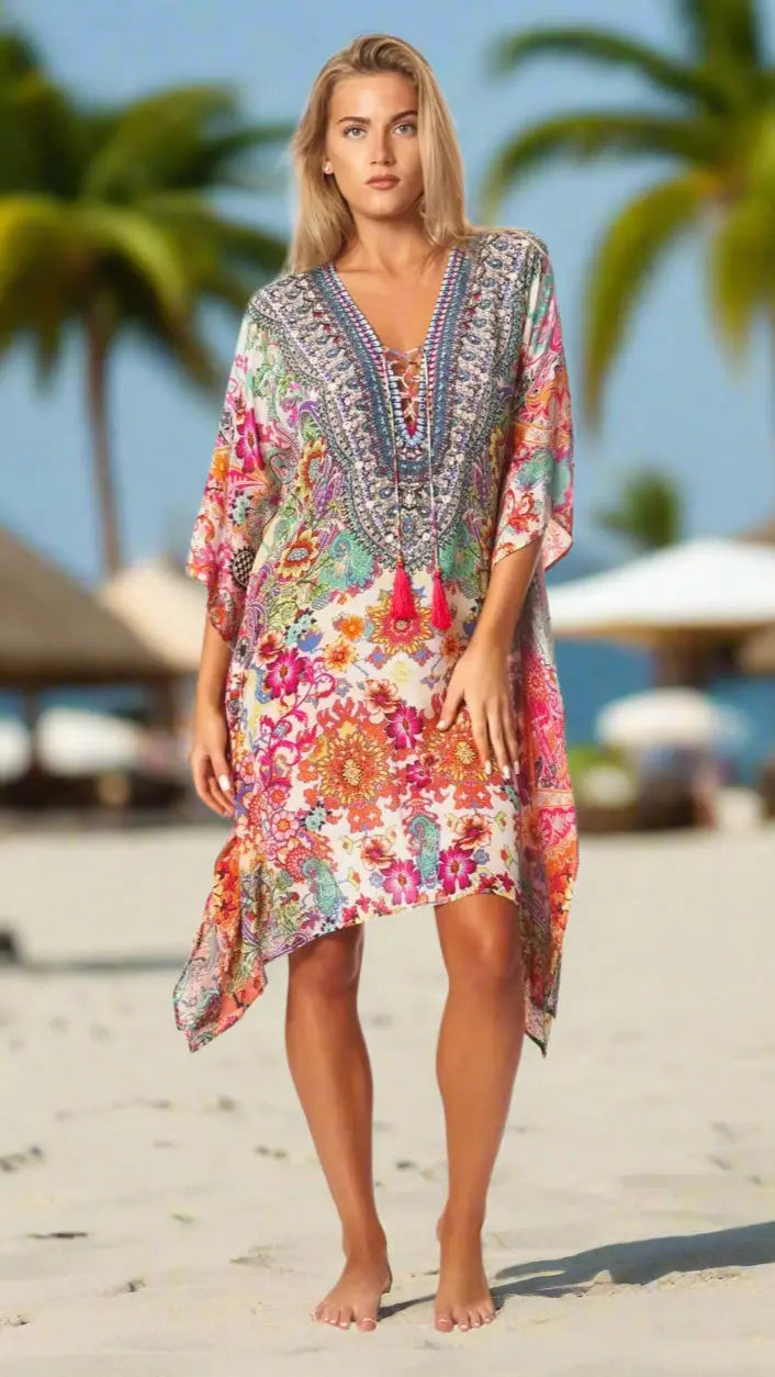 A woman stands barefoot on a sandy beach in a La Moda Clothings Short Kaftan Dress for Women, featuring vibrant pink, red, green, and blue patterns. Palm trees and blurred beach umbrellas complete the relaxed, summery vibe.