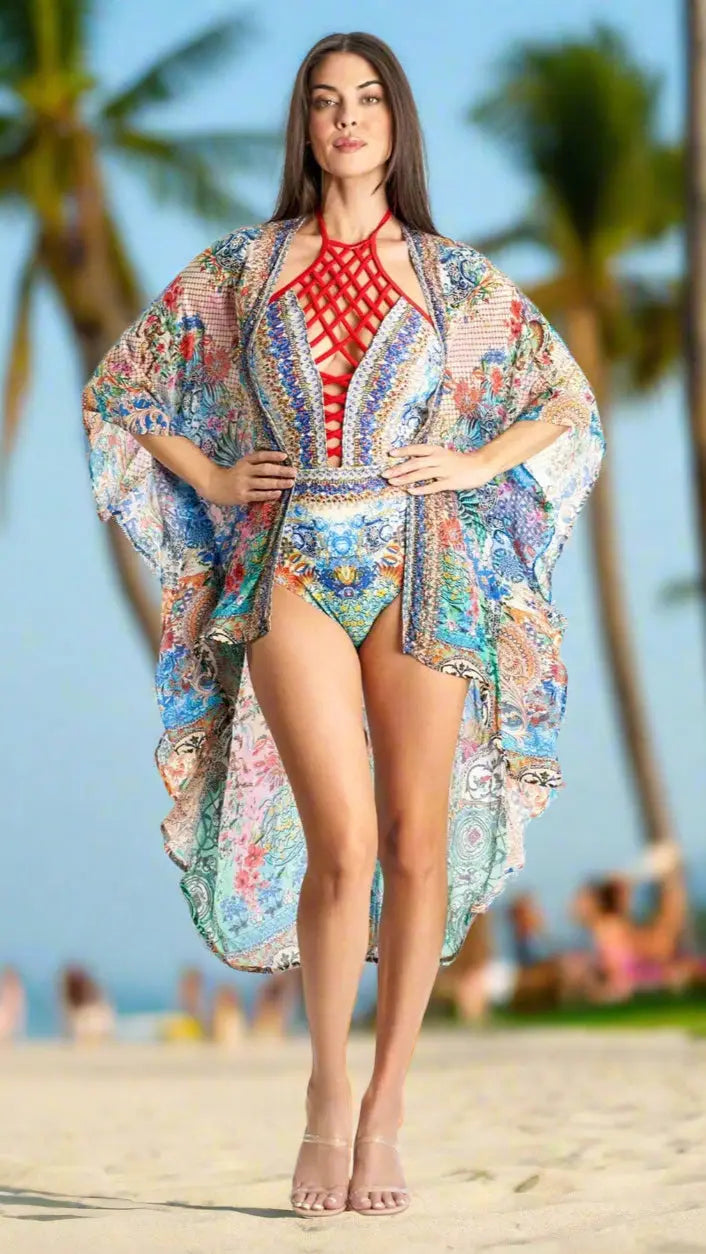 A woman stands confidently on the beach in a vibrant patterned swimsuit and the Kimono Cover Up in Viscose Silk by La Moda Clothings. She wears clear heels, has long brown hair, neutral makeup, with palm trees and the sea in the background.