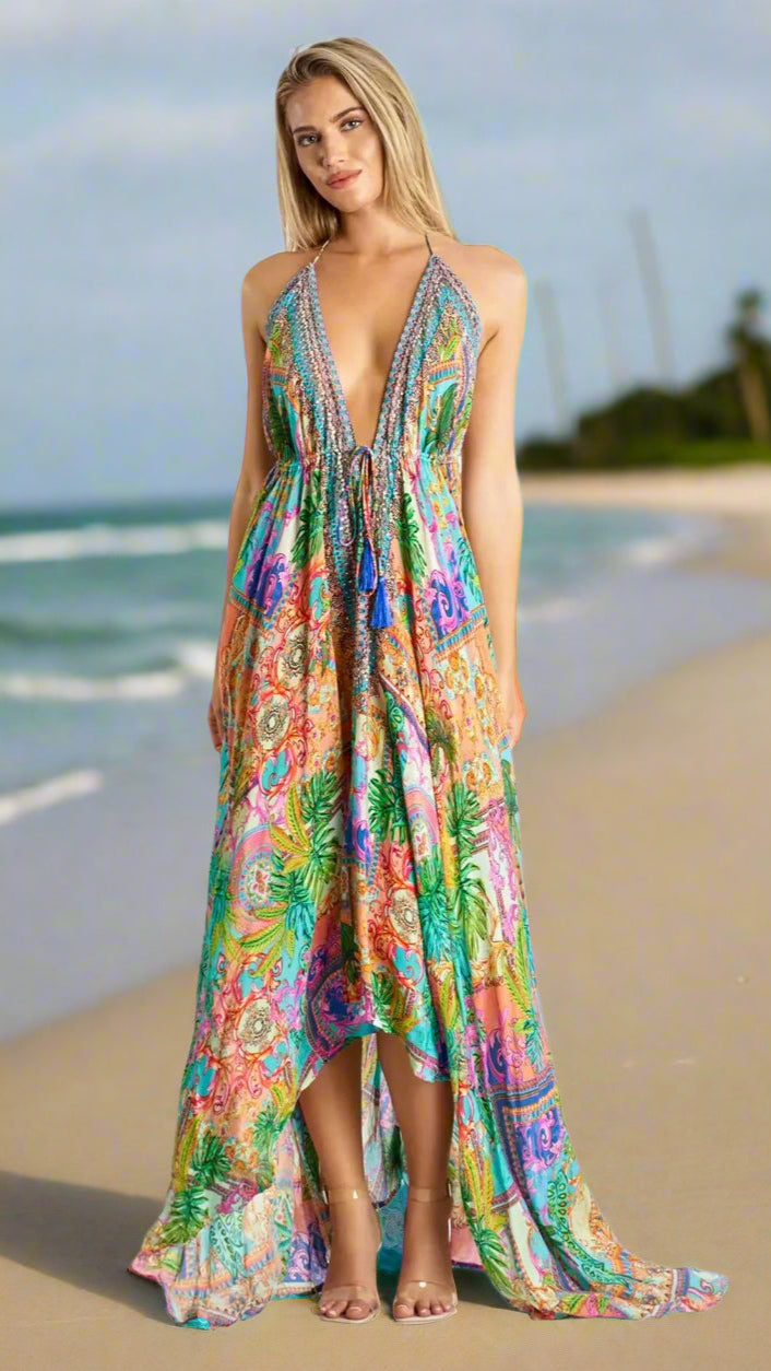 A woman with light hair stands barefoot on a sandy beach, wearing the Lamodaclothing Designer Halter Neck Maxi Dress. The dress features a deep V neckline, embroidered waist, and vibrant floral and paisley patterns, with the ocean and sky behind her.