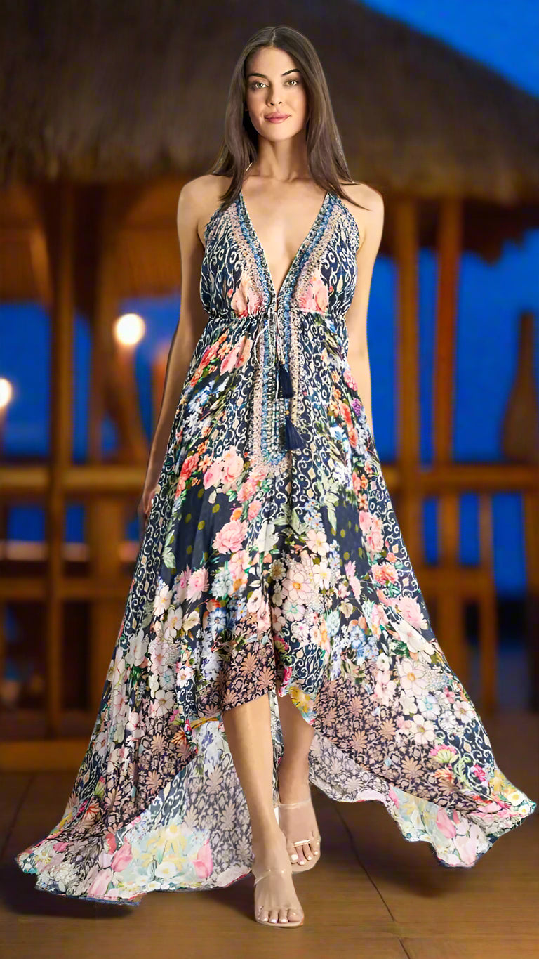 A woman with long brown hair models the La Moda Clothing Resort Wear Halter Maxi Dress with a plunge neckline, featuring blue and white geometric and floral prints. She stands barefoot on a wooden floor before a tropical, thatched-roof backdrop.