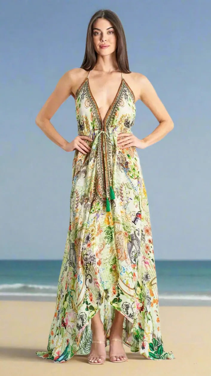 A woman stands on a sandy beach with the ocean behind her, wearing La Moda Clothing’s Halter High Low Maxi Resort Dress for Summer, featuring a plunging neckline, colorful florals, tie waist detail, and nude heels. Her arms rest on her hips.