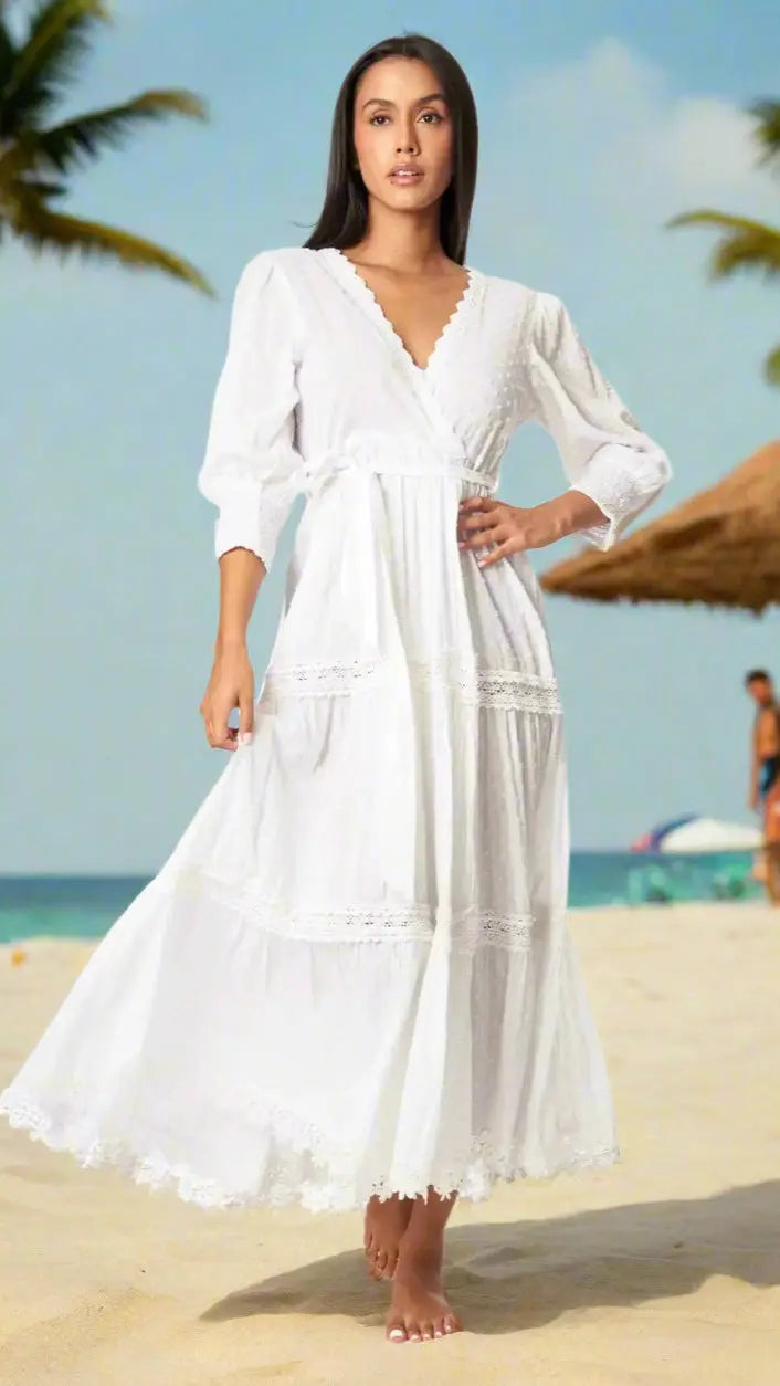 A woman stands barefoot on a sandy beach under palm trees and a thatched umbrella, wearing a La Moda Clothing Wrap Women’s Maxi Dress for Summer with lace trim and three-quarter sleeves—a perfect look for tropical days.