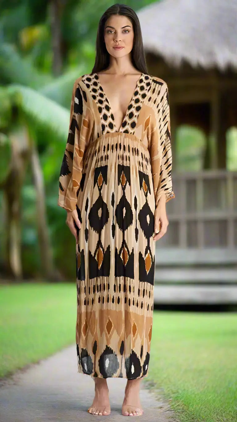 A woman with long dark hair stands barefoot on a path near a thatched-roof hut, wearing La Moda Clothing's Summer Maxi Dress—a flowy, long-sleeved dress with a deep V-neck and geometric tan, black, and cream pattern.