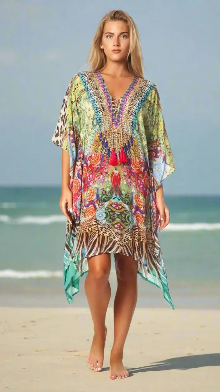 A woman with long blonde hair walks barefoot on a sandy beach, wearing a vibrant Animal Print Kaftan by La Moda Clothings. The kaftan features multicolored patterns, including animal prints and florals, with the ocean softly blurred behind her.