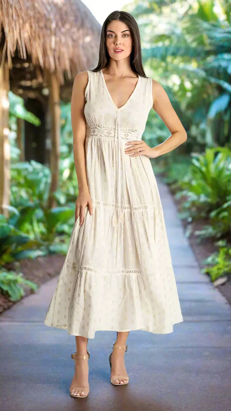 A woman stands on a garden path wearing the Azalea Tiered Maxi Resort Dress for Island Women by La Moda Clothing. The sleeveless, cream ankle-length dress features subtle embroidery. Lush greenery and a thatched-roof structure are in the background.