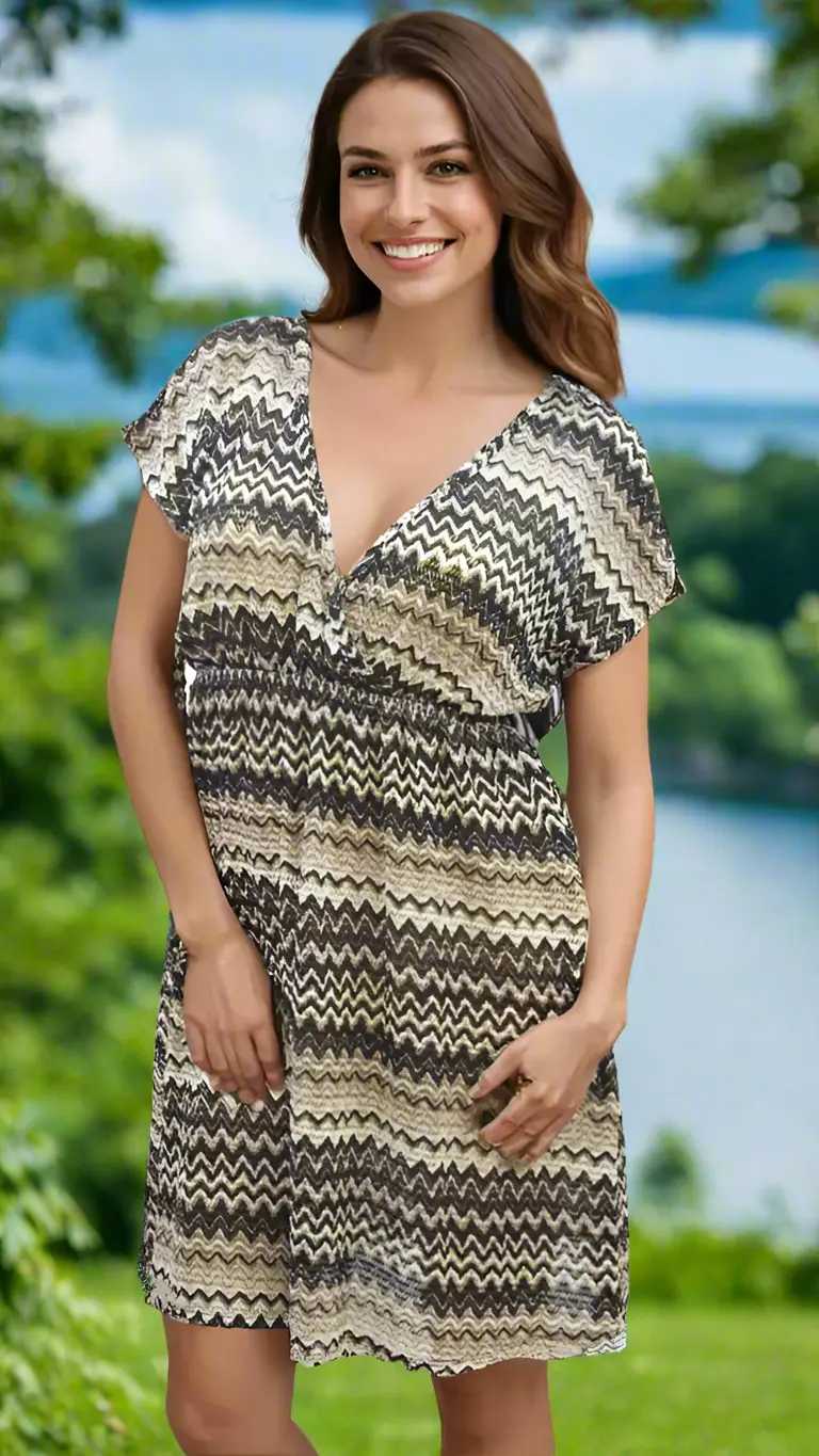 A woman with long brown hair smiles outdoors by greenery and a lake, wearing the La Moda Clothing Beach Cover Up Dress—a short-sleeved, knee-length dress featuring a black, beige, and cream zigzag pattern.