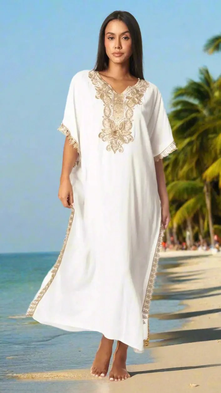 A woman with long dark hair stands barefoot on a sandy beach, wearing the La Moda Clothing Beach Maxi Kaftan Dress for Women, featuring gold embroidery. Palm trees and a calm blue sea are in the background under a clear sky.
