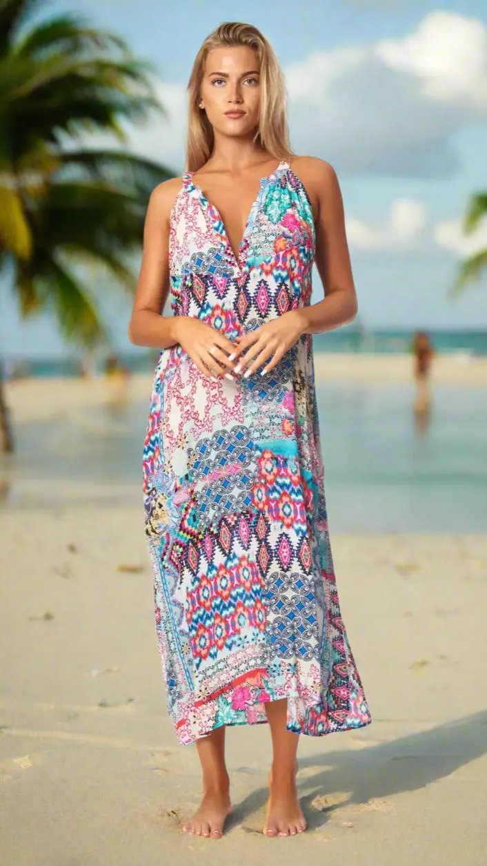 A woman with long blonde hair stands barefoot on a sandy beach in the Beach Resort Vacation Dress by La Moda Clothing—a colorful, patterned sleeveless dress with a deep V-neck. Palm trees and blurred figures appear in the sunny background.