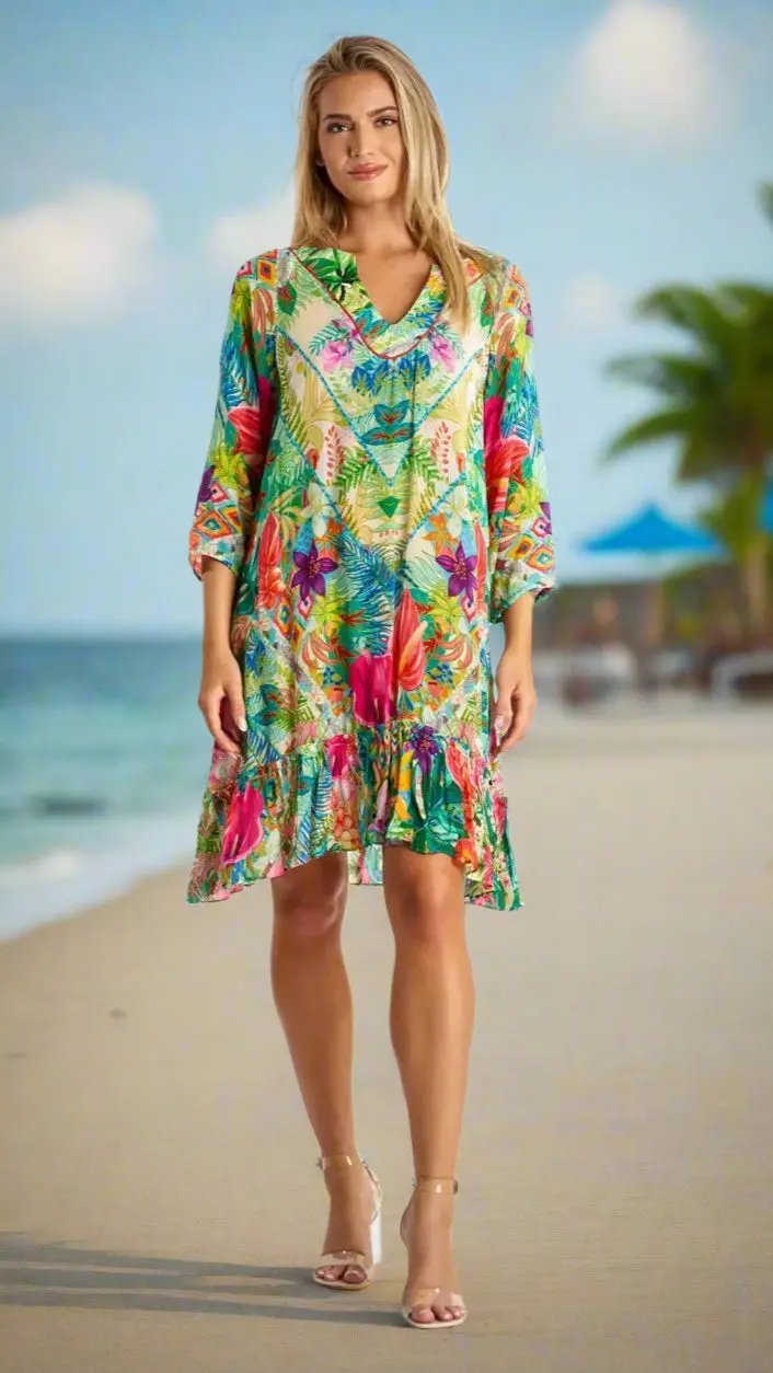 A woman stands on a tropical beach, smiling in the La Moda Clothings Bohemian Summer Dress for Beach & Beyond, featuring three-quarter sleeves and a ruffled hem. Palm trees and ocean waves blur behind her under a blue sky.