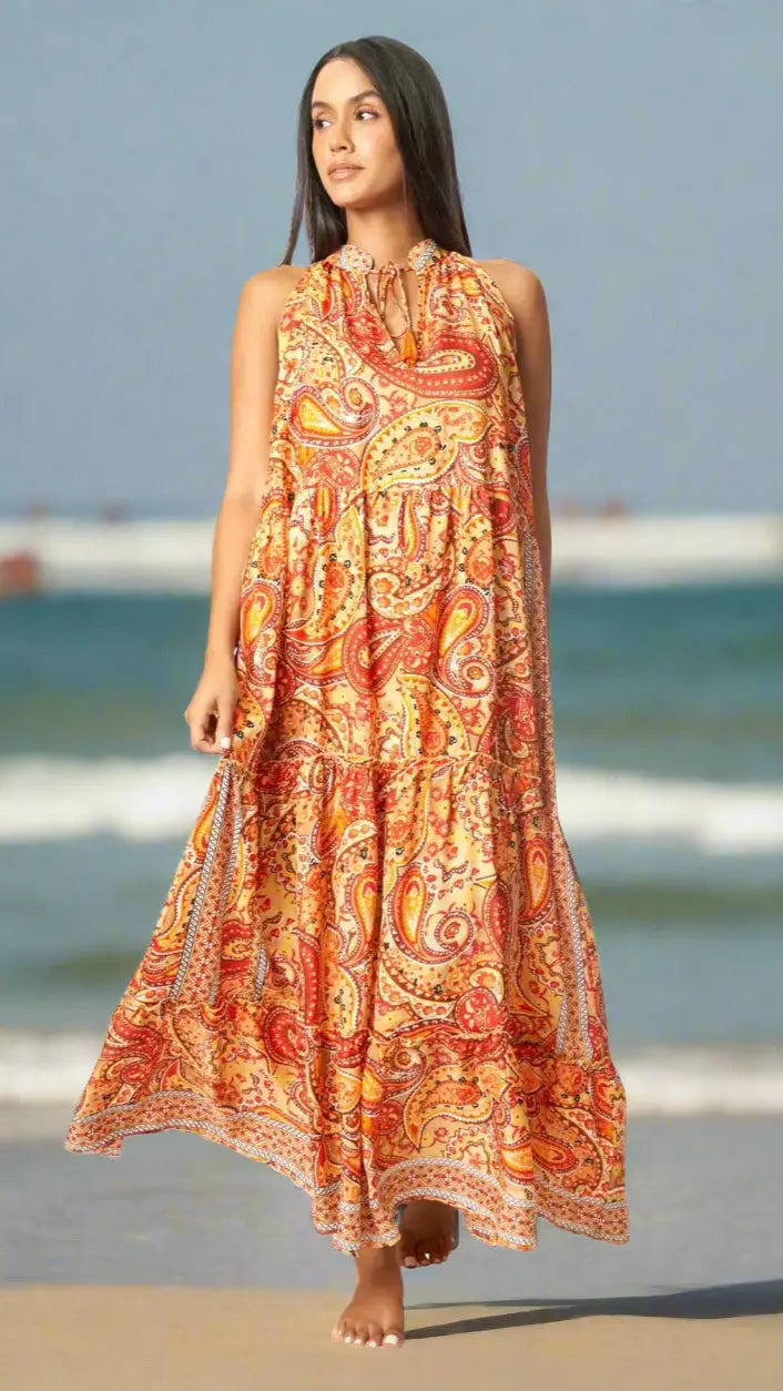 A woman with long dark hair stands barefoot on the beach in a La Moda Clothing Boho Positano Maxi Dress, featuring a bright orange, yellow, and red paisley print—perfect for Caribbean vacations—with ocean waves and blue sky behind her.