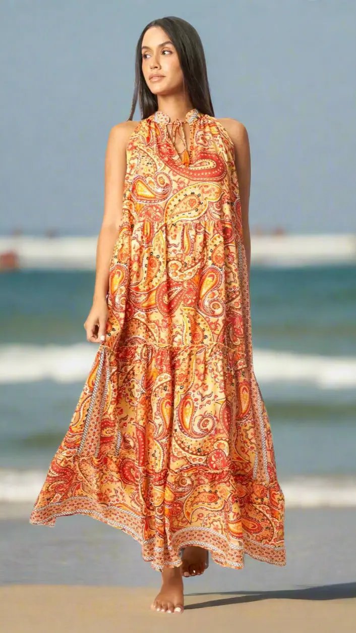 A woman with long dark hair stands barefoot on the beach in a La Moda Clothing Boho Positano Maxi Dress, featuring a bright orange, yellow, and red paisley print—perfect for Caribbean vacations—with ocean waves and blue sky behind her.