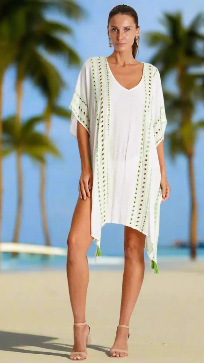 A woman stands on a sandy beach with palm trees and blue sky, wearing the La Moda Clothing Caftan—a white, loose V-neck cover-up with green detailing and tassels. Her hair is pulled back and she looks ahead with a neutral expression.