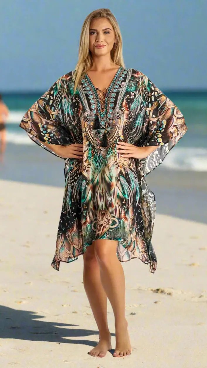 A woman stands barefoot on a sandy beach, smiling and wearing the Lamodaclothing Caftan Light weight Lounger Dress featuring wide sleeves with teal, brown, black, and beige patterns. The ocean and blue sky form the backdrop.