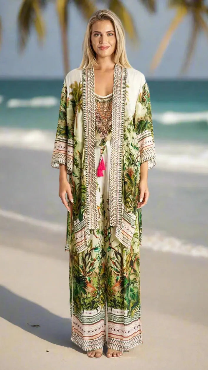A woman stands barefoot on the beach in a La Moda Clothings Cardigan Kimono Cape for Summer Wear with green leafy and pink tropical patterns. Palm trees and the ocean are behind her as she smiles, her long blonde hair loose over her shoulders.