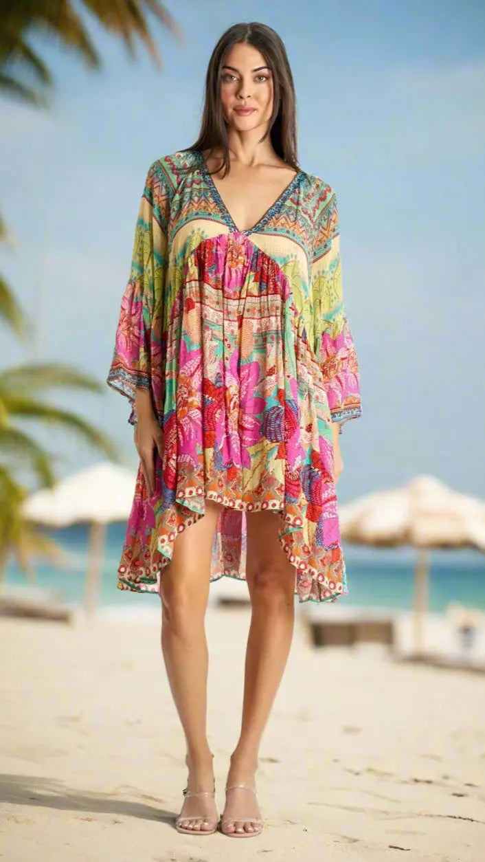 A woman stands barefoot on a sandy beach wearing the La Moda Clothing Casual Resort Mini Dress, a colorful, loose-fitting floral summer trend with wide sleeves. Palm trees and white umbrellas blur in the bright, sunny background.