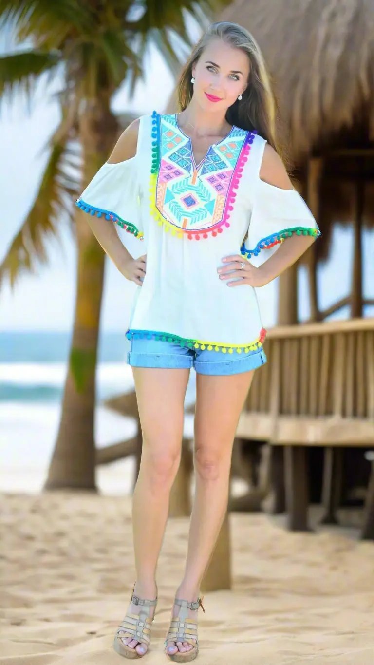 A woman smiles barefoot on the sand, wearing denim shorts and a lamodaclothings Club Moda Beach Top with playful pompoms. Palm trees and a thatched hut are in the background, and her long hair flows freely.