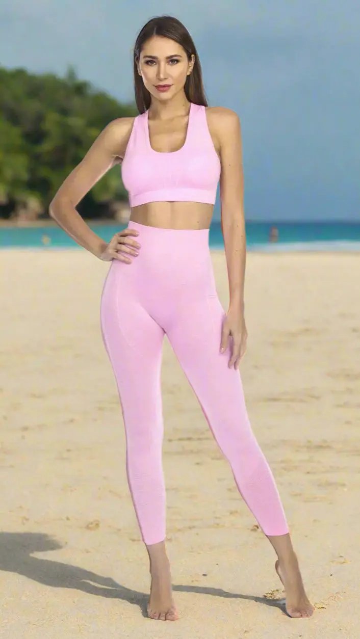 A woman with long brown hair and light skin stands barefoot on a sandy beach in the Club Moda Textured Scoop Neck Seamless Sports Bra by La Moda Clothings and high-waisted leggings, framed by green trees and blue ocean beneath a clear sky.