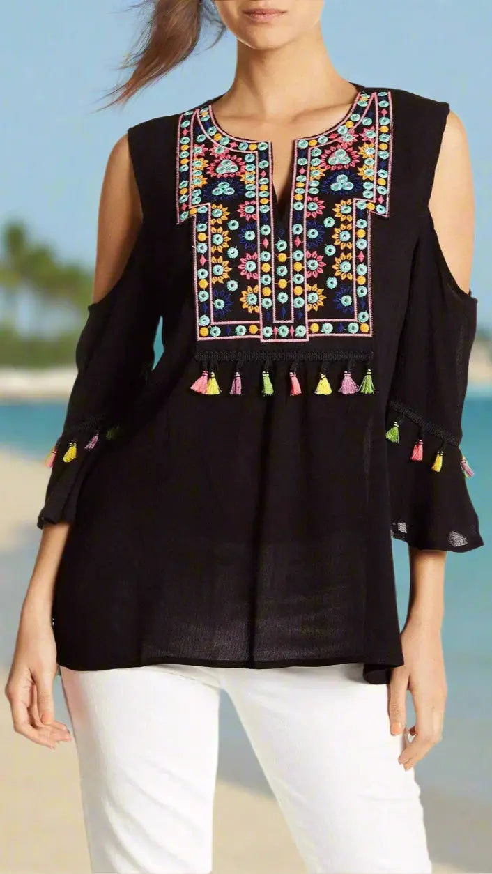 A woman wears the lamodaclothings Cold Shoulder Beach Top with Colorful Tassels, paired with white pants, standing on a beach with blurred palm trees and blue sky in the background.