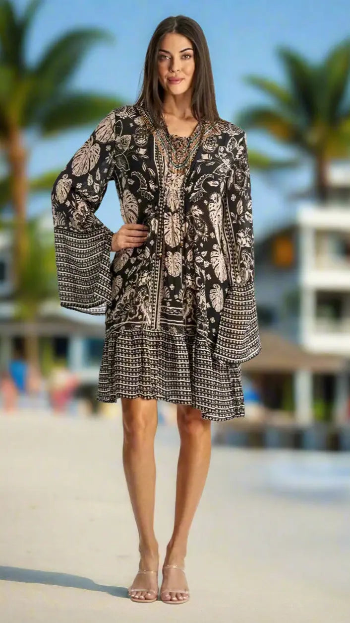 A woman with long brown hair stands outdoors in the sun wearing the La Moda Clothing Comfy Resort Wear Short Mini Dress featuring beige floral and geometric patterns, flared sleeves, a knee-length skirt, and clear open-toe heels. Palm trees are behind her.