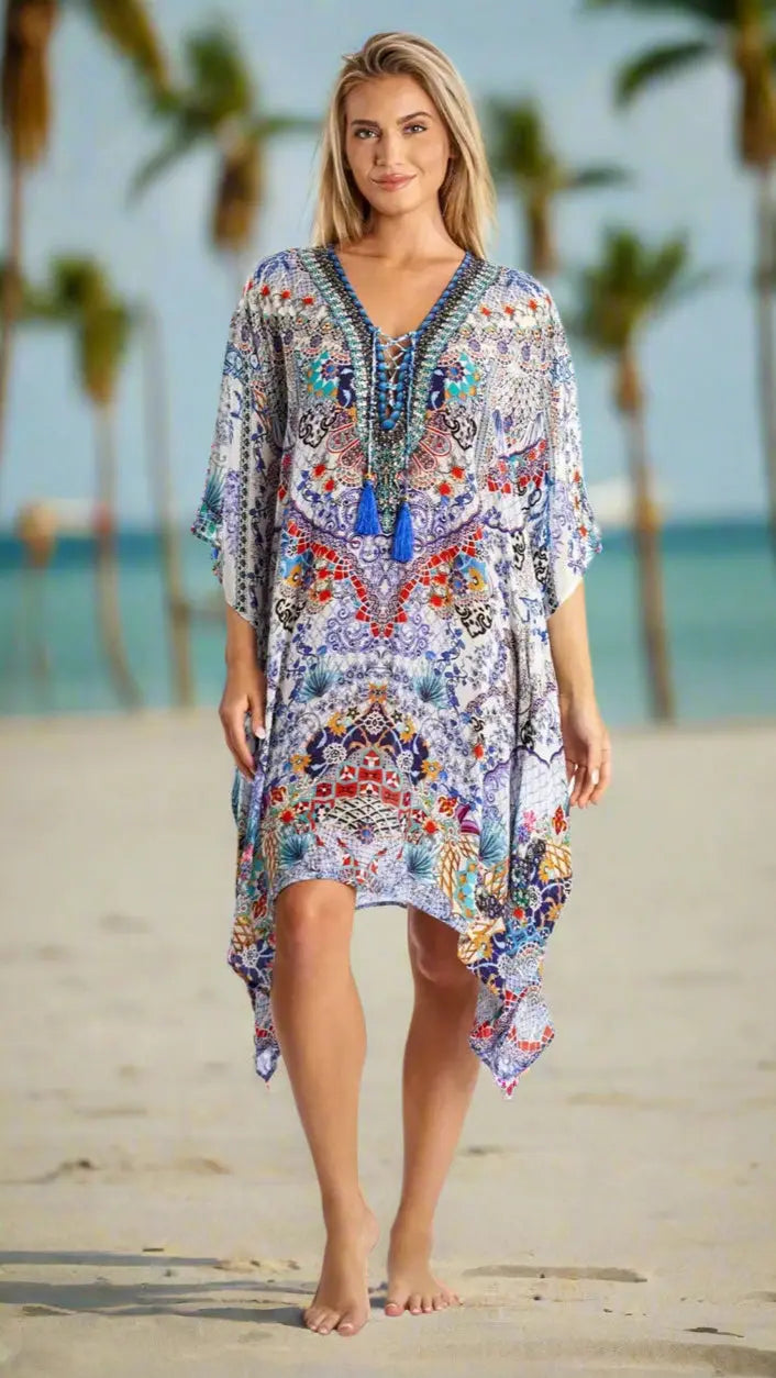 A woman stands barefoot on a sandy beach with palm trees and blue ocean, wearing La Moda Clothings’ Designer Caftans Kaftan for Tropical Destinations—a flowing, patterned V-neck dress in blue, red, and orange with a tassel necklace.