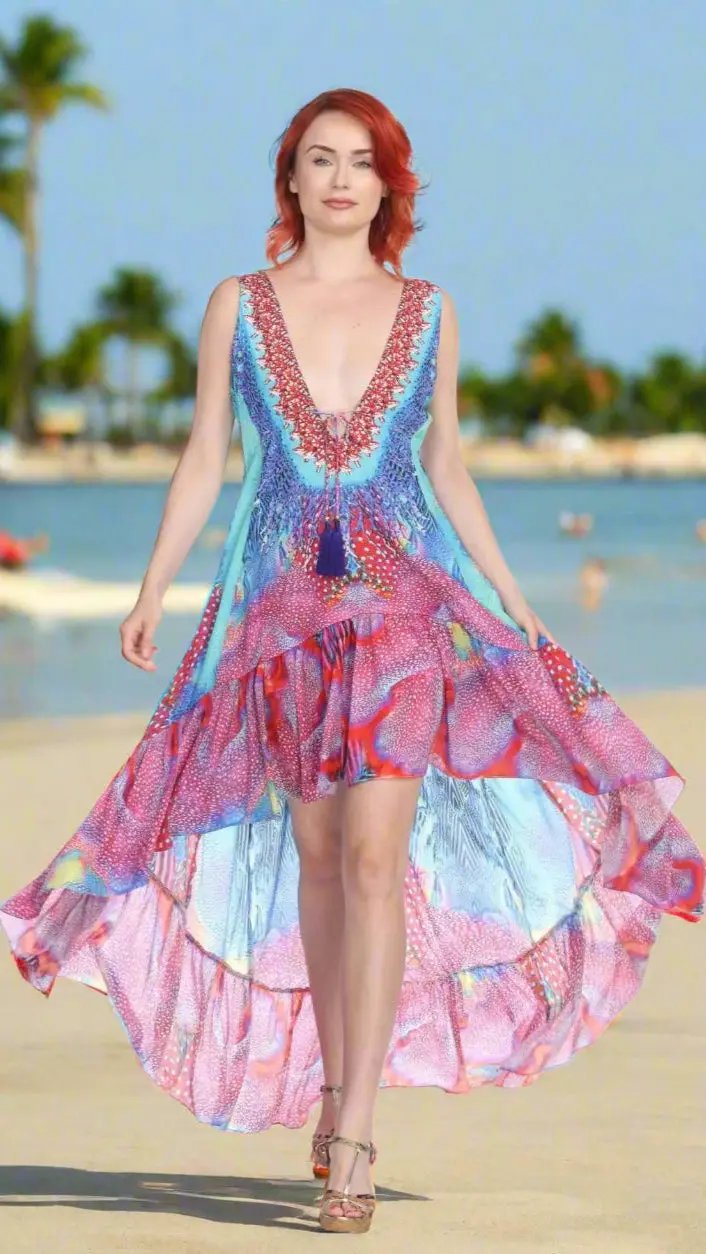 A red-haired woman strolls confidently on a sandy beach in the Lamodaclothing Designer High Low Multi-Color Vacay Dress. Palm trees and the ocean blur in the background beneath a clear sky.