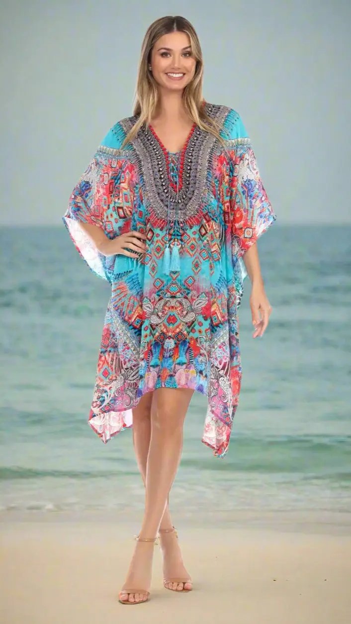 A smiling woman stands on a beach in a La Moda Clothings Designer Silk Kaftan, featuring vibrant blue, red, and orange geometric and floral patterns. She pairs it with beige strappy sandals, as the ocean and sky create a serene backdrop.