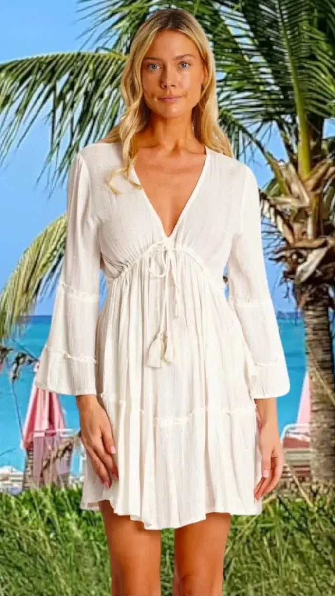 A woman with long blonde hair wears the La Moda Clothing Dyed Mini Dress, featuring a white, long-sleeved v-neck design with a waist tie. She stands by palm trees and pink beach umbrellas, with the ocean under a clear sky in the background.