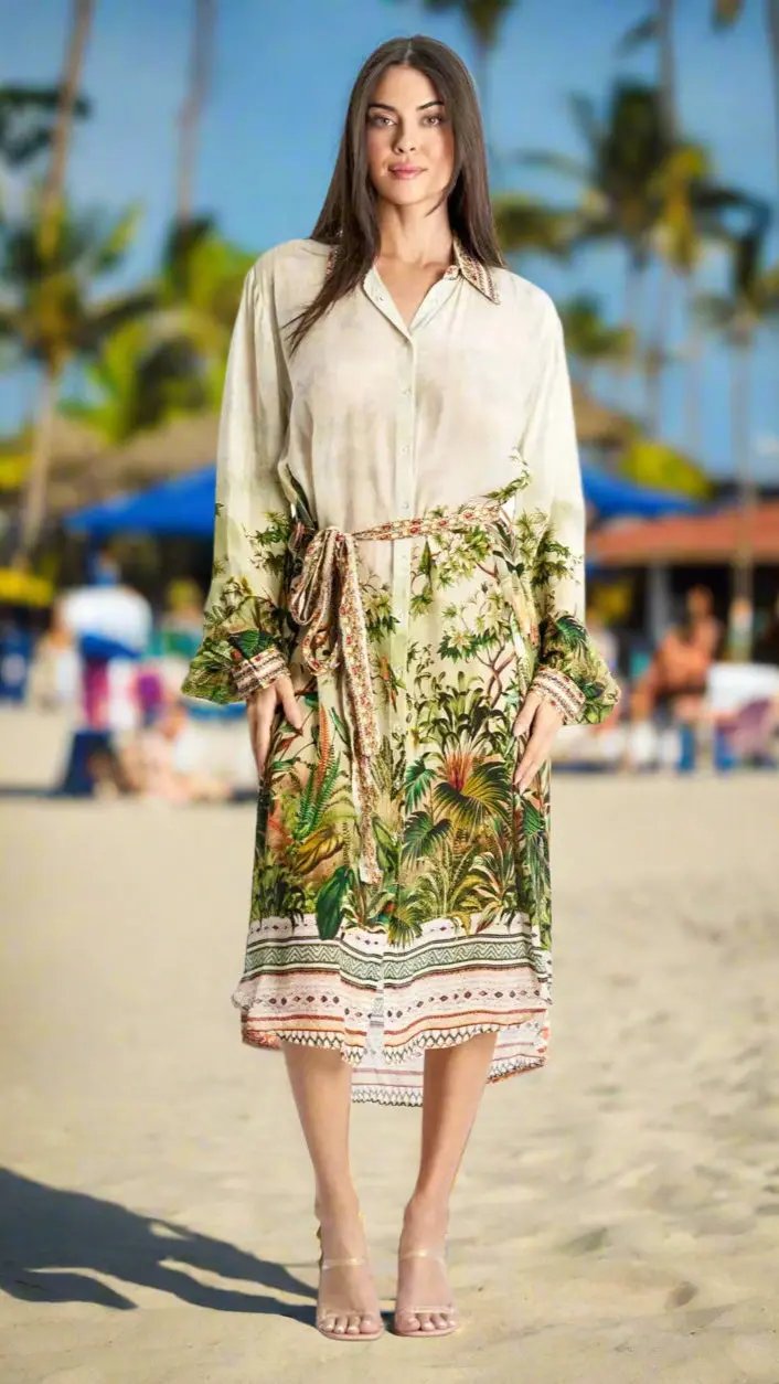 A woman stands barefoot on the sand in a cream tropical-print Easy Shirtdress Cover-Up Resortwear by La Moda Clothings. She smiles gently at the camera, with palm trees and blue umbrellas in the sunny background.