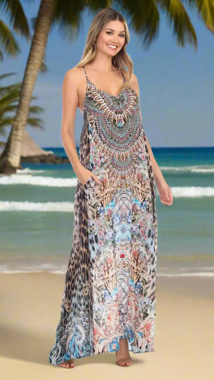 A woman smiles barefoot on a tropical beach, wearing the Elegant Maxi Pocket Dress in Viscose Silk by La Moda Clothings—an animal-print, intricately designed dress with thin straps and pockets. Palm trees, ocean, and a hut blur in the background.