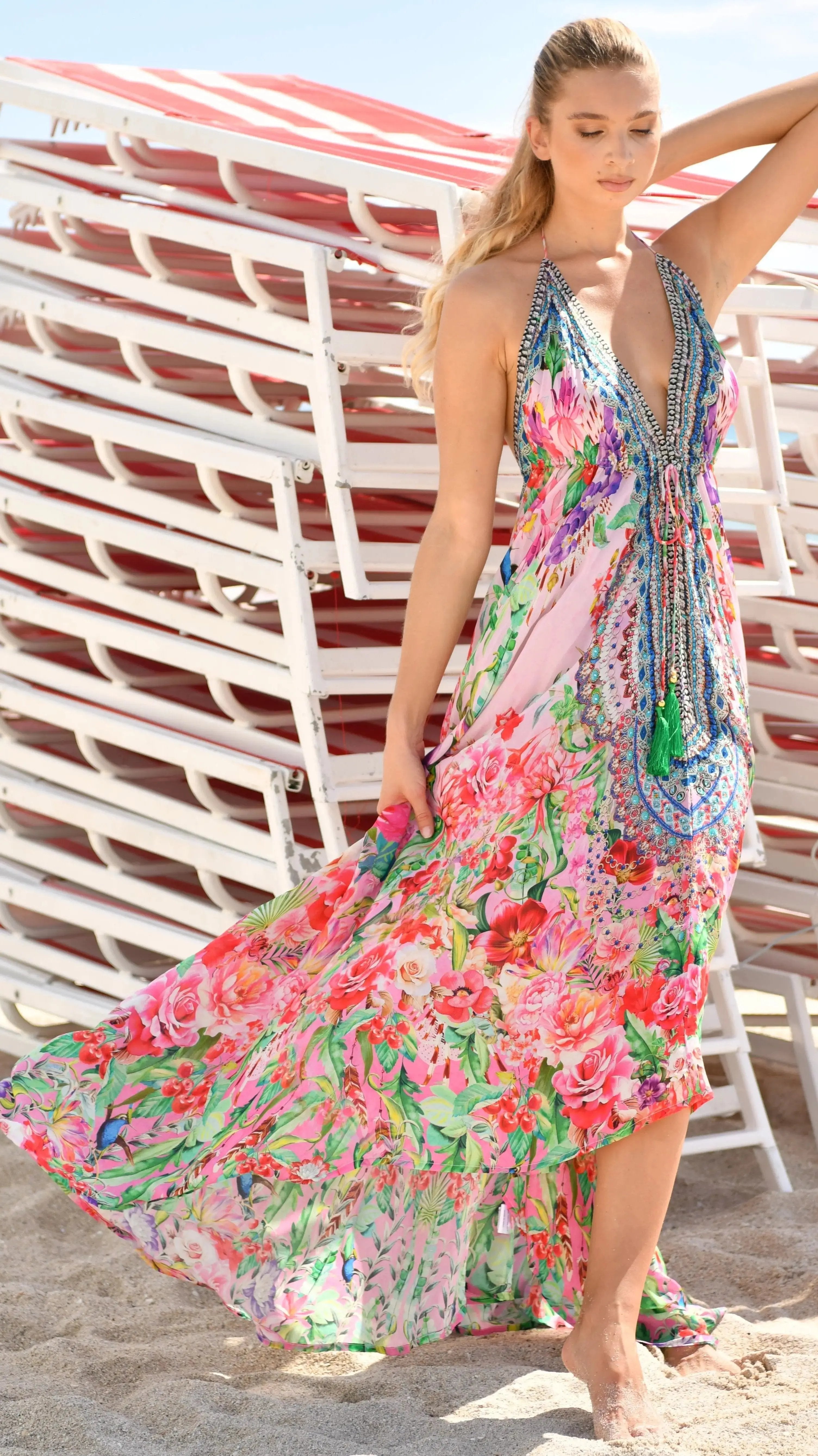 A woman with light hair stands on sand before white and red lounge chairs, wearing Lamodaclothing’s Endless Summer Designer Halter Neck Backless Resort Dress in a vibrant pink, green, and blue floral print. She poses with one arm raised above her head.