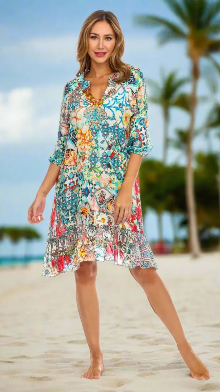 A woman with wavy blonde hair stands barefoot on a sandy beach, wearing the La Moda Clothings Ethnic Style Summer Dress featuring colorful floral and geometric prints. Palm trees and a blurred tropical sky complete the scene.