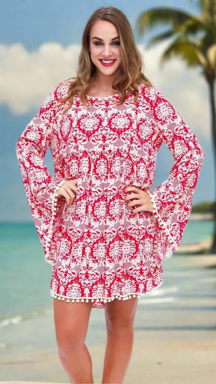 A woman smiles on a beach by a palm tree, wearing lamodaclothings' Full Sleeved Beach Cover Up Dress—a red and white long-sleeve piece with flared sleeves and white pom-pom trim. The ocean and clouds form the scenic backdrop.