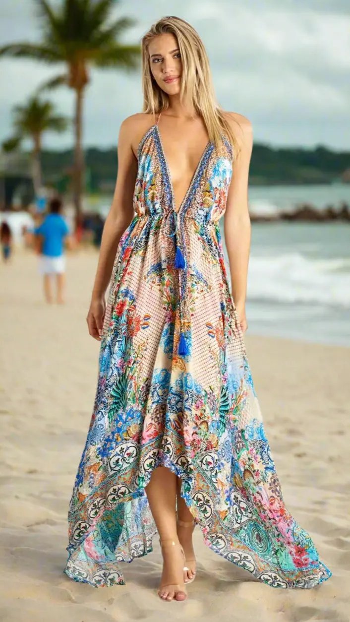 A woman stands barefoot on a sandy beach wearing the Halter Maxi Resort Dress by La Moda Clothing, featuring a colorful, flowy design. Palm trees and waves are in the background as people walk along the shore under a cloudy sky.