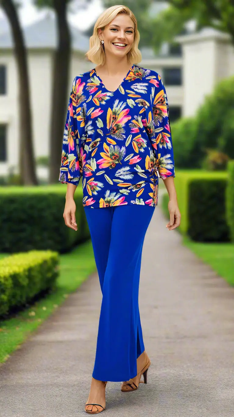 A smiling woman strolls a garden path in La Moda Clothing's Knit Bell Sleeve Resort Wear Tunic Top, featuring a blue floral print with orange, yellow, and pink flowers. Blurred trees and a white house appear in the background.