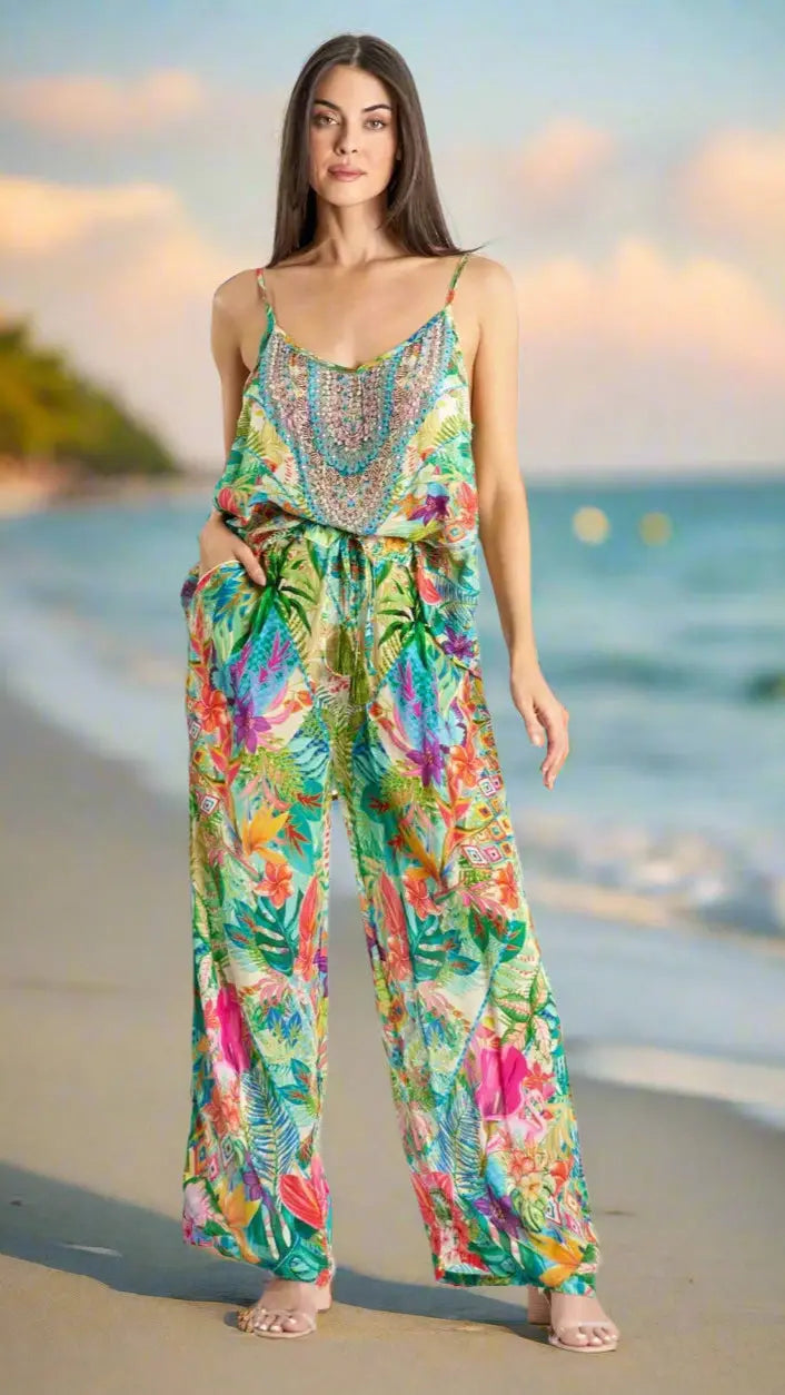 A woman stands barefoot on a sandy beach at sunset, wearing the La Moda Resort Lifestyle Pant Set by La Moda Clothing. Her straight brown hair is loose, and the ocean with blurred trees appears in the background.