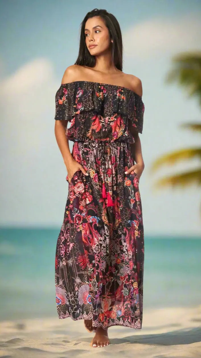 A woman with long dark hair stands barefoot on a sandy beach, wearing the La Moda Clothings Long Off the Shoulder Dress featuring a vibrant floral and paisley print. She looks to the side, with a palm tree and ocean blurred in the background.