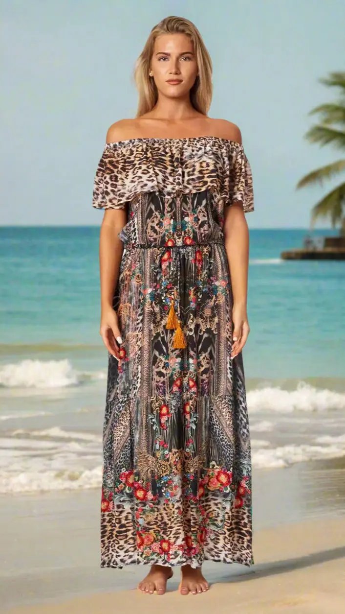 A woman stands barefoot on a sandy beach in the Long Off the Shoulder Dress by La Moda Clothings, featuring a leopard print top and a colorful floral skirt. Gentle waves, a palm tree, and a calm sea complete the serene background.