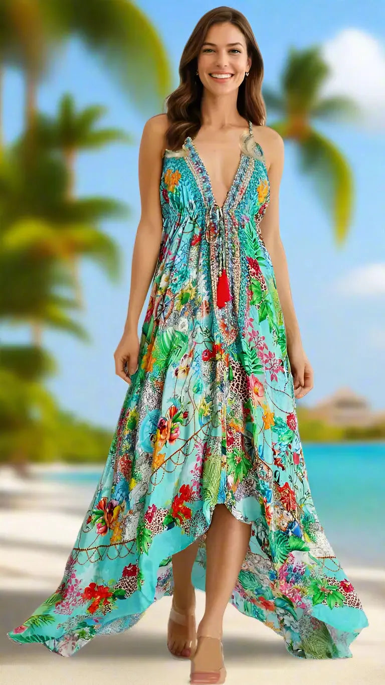 A smiling woman strolls a tropical beach in the La Moda Clothing Luxury Maxi Resort Dress, featuring a colorful, flowy design with a plunging neckline and floral patterns, palm trees and blue ocean softly blurred behind her.