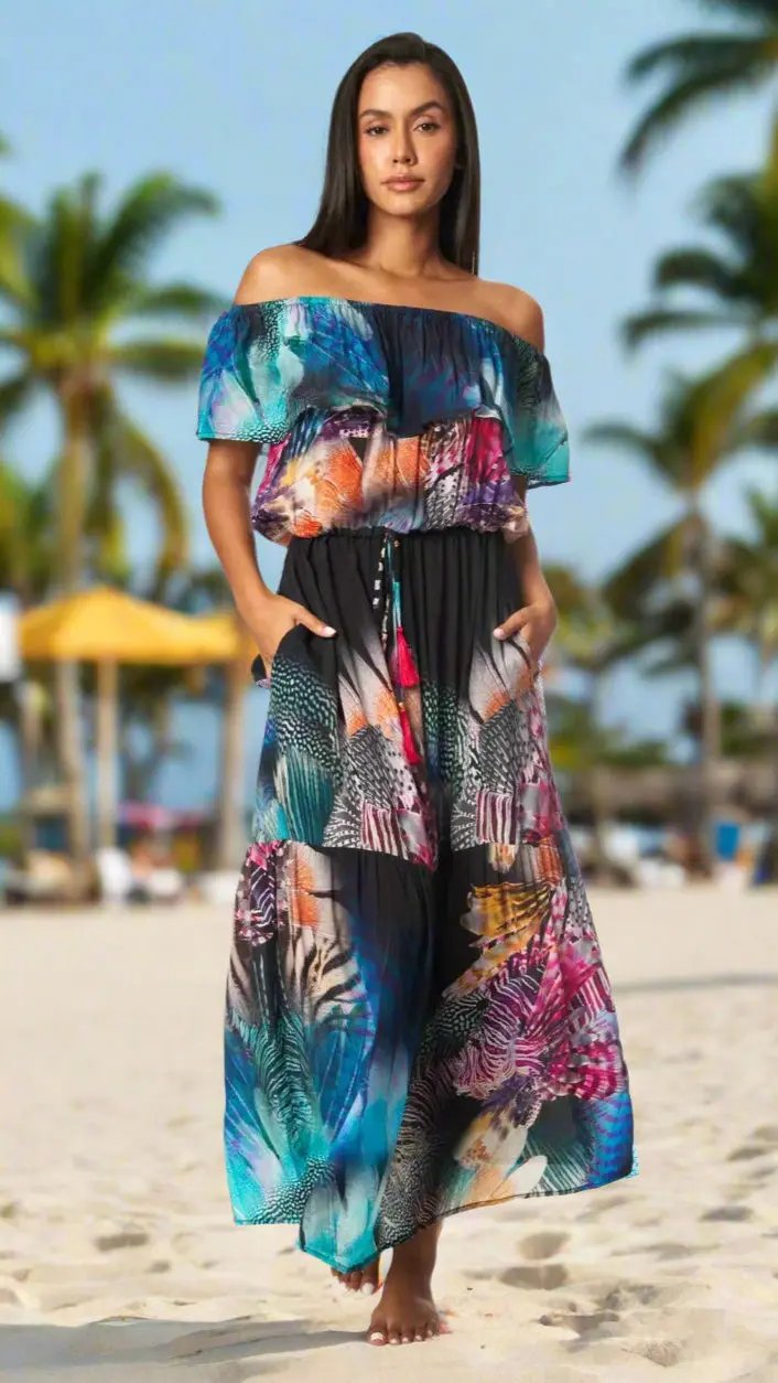 A woman stands barefoot on a sandy beach, wearing the Maxi Long Off the Shoulder Dress by La Moda Clothings, featuring colorful tropical leaf and feather patterns. Palm trees and blurred umbrellas in the background evoke a warm, tropical vibe.