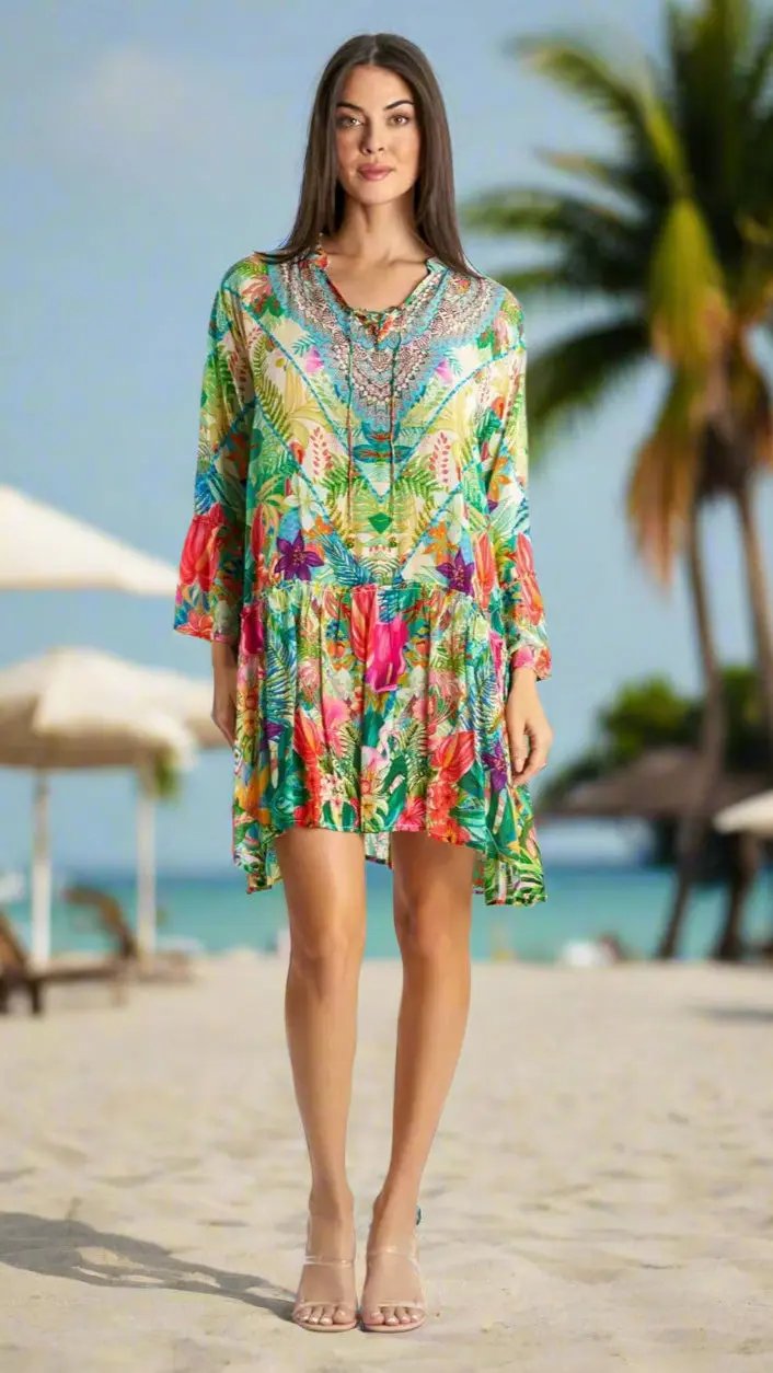 A woman with long brown hair smiles softly as she stands barefoot on a sunny beach, wearing the La Moda Clothing Mini Dress for Beach Resort & Cruise. The brightly colored, floral-patterned dress with long sleeves contrasts against blurred palms and lounge chairs.