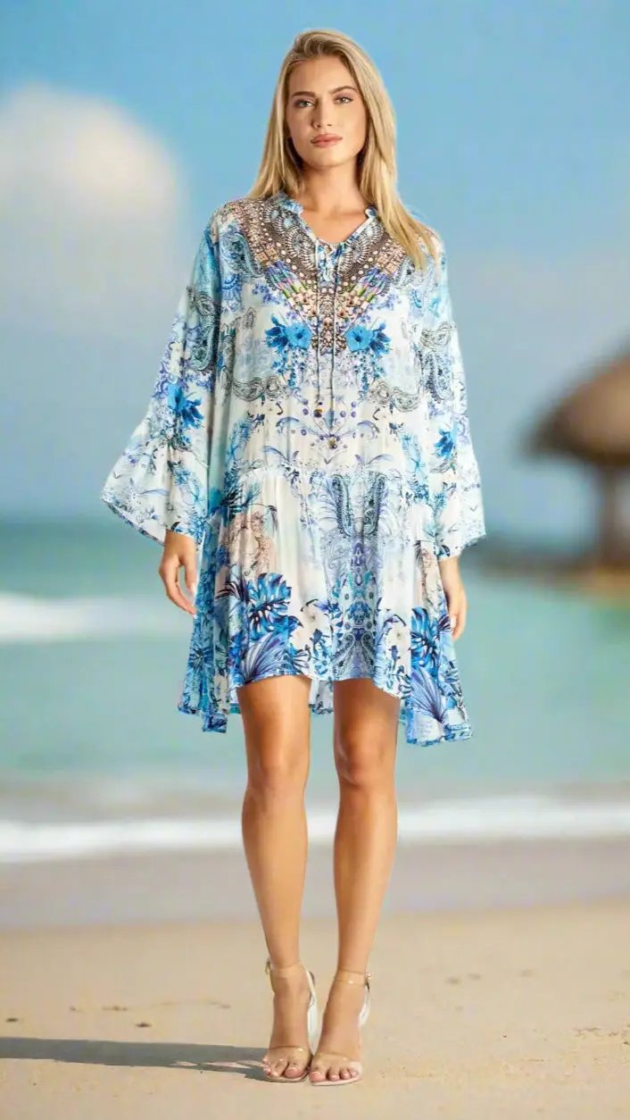 A woman with straight blonde hair stands on a sandy beach wearing La Moda Clothing’s Mini Dresses for Beach Resort & Cruise in a blue and white floral paisley print, paired with nude strappy heels. The sea and a thatched-roof hut blur in the background.
