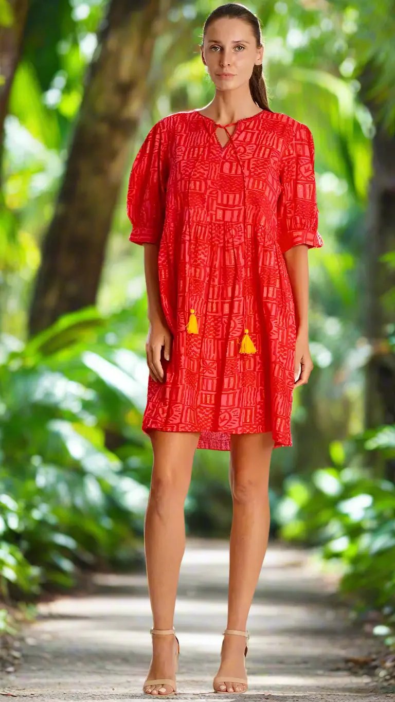 A woman stands on a sunlit path wearing the La Moda Clothing Mini Summer Dress, featuring a loose fit with geometric patterns, yellow tassels, and puffed half-sleeves, paired with beige strappy heels. Her hair is pulled back and she faces forward.
