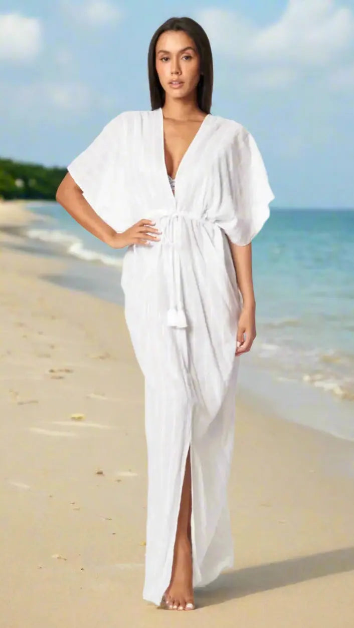 A woman stands barefoot on a sandy beach, wearing the lamodaclothings Moda Long Kaftan-Style White Beachwear Cover Up with short sleeves and a waist tie. The ocean and a blue sky with clouds are behind her as she looks at the camera, relaxed.