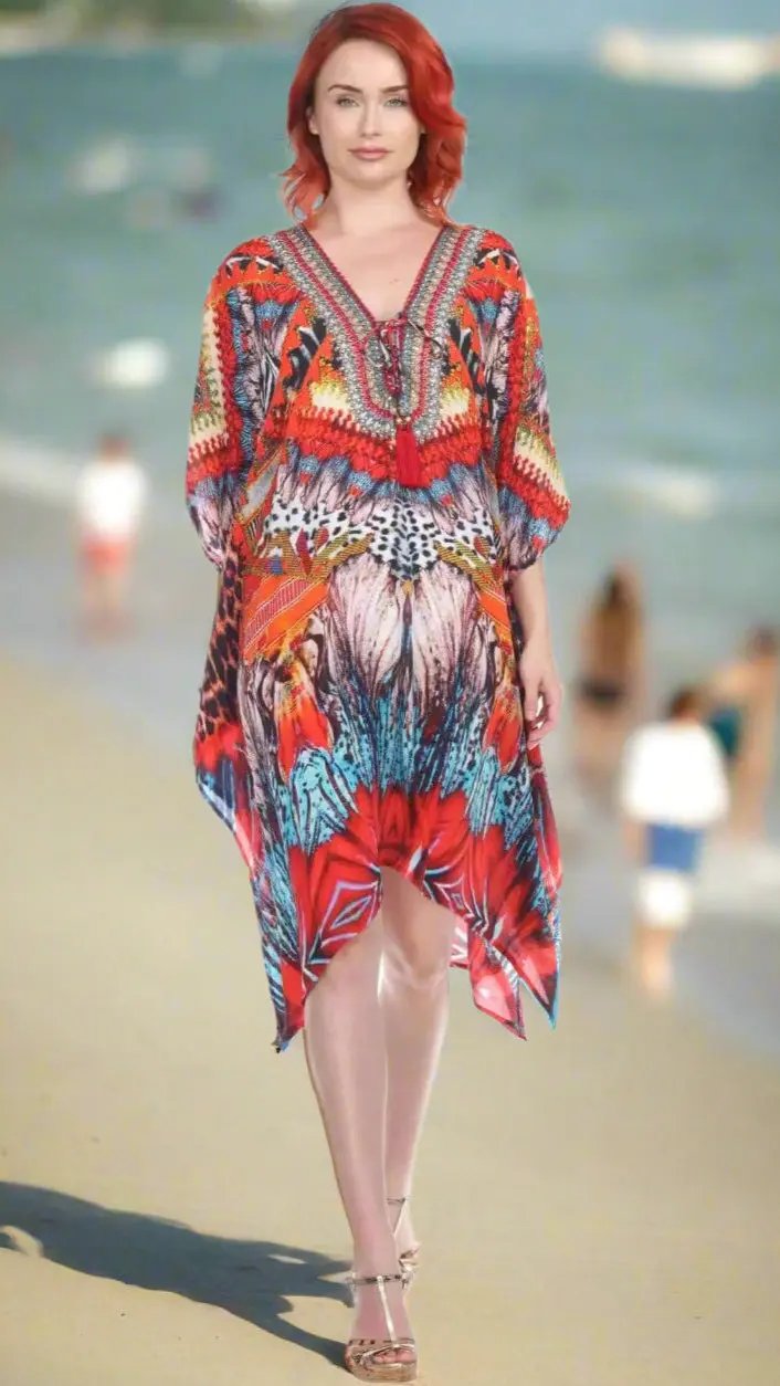 A woman with red hair and light skin walks on the beach in a Lamodaclothing Multi-Color Trendy Vacation Beach Caftan Dress, featuring vibrant patterns. She wears sandals as blurred beachgoers and the sea appear in the bright background.