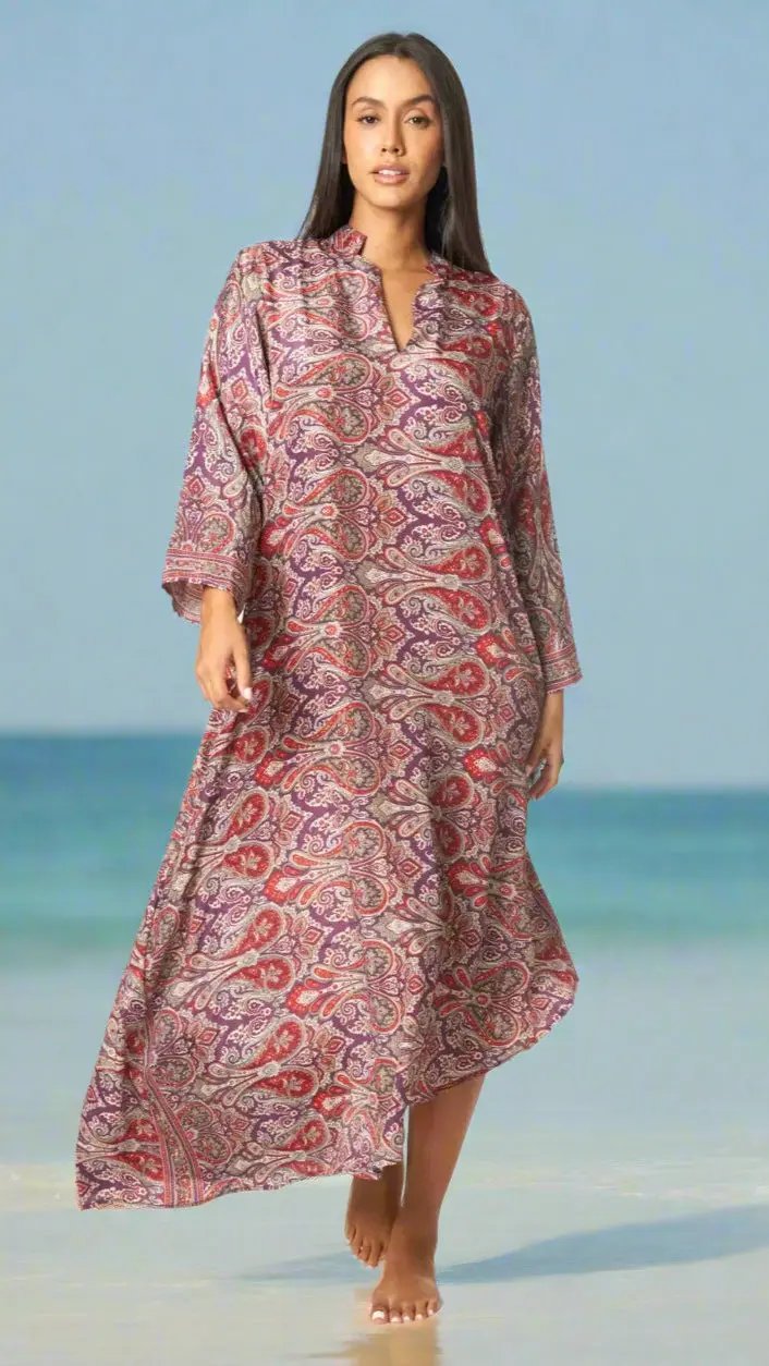A woman with long dark hair walks barefoot on the beach in La Moda Clothing's Positano Maxi Caftan Dress for Spring Summer, featuring a red and purple paisley pattern. The serene sea and blue sky create a calm, summery vibe.