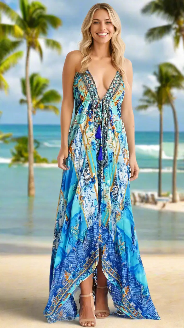 A smiling woman with long blonde hair wears the Resort Maxi Dress by La Moda Clothing, a flowy blue patterned V-neck design with tassels, as she stands on a sandy beach with palm trees and ocean waves under a partly cloudy sky.