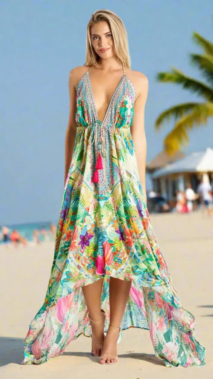 A woman with blonde hair models the La Moda Clothings Resort Wear Maxi Dress for Summer Vacations, a colorful, tropical-print design with a deep V-neck and high-low hem, standing barefoot on a beach with palm trees under a clear blue sky.