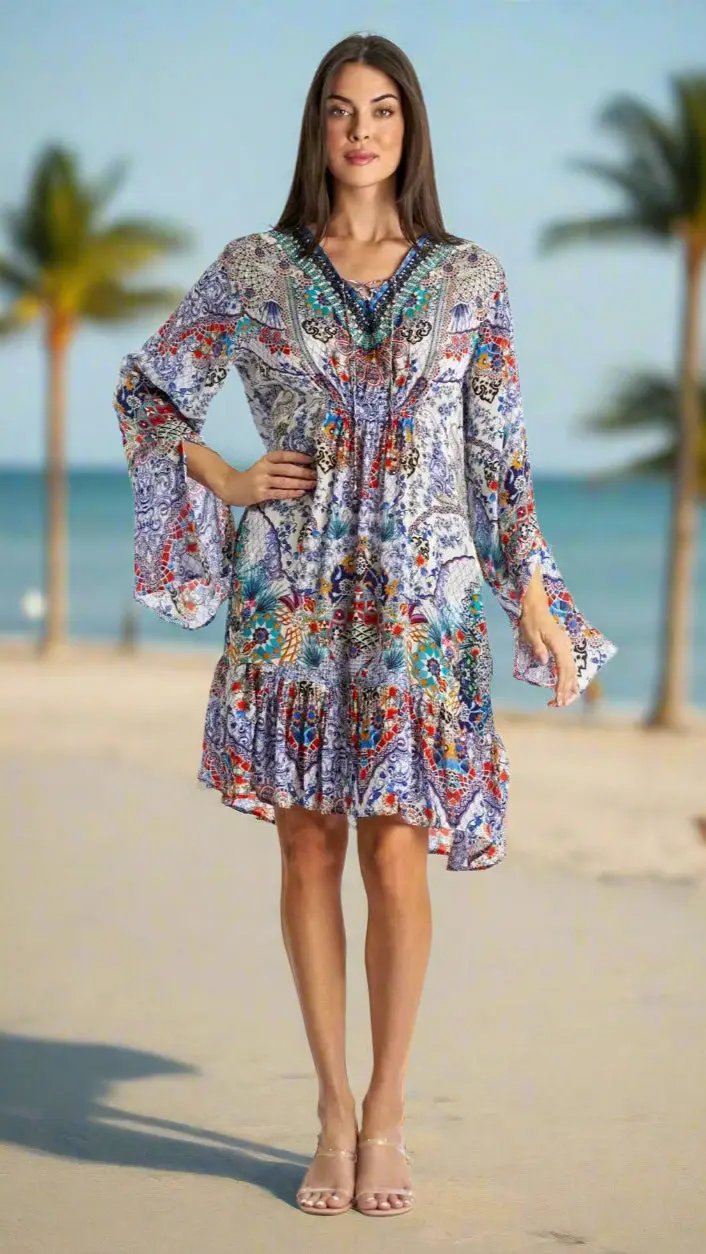 A woman with long brown hair smiles on a sandy beach, wearing La Moda Clothing's Resort Wear Short Mini Dress in trendy prints and clear heeled sandals, with palm trees and the ocean in the background.